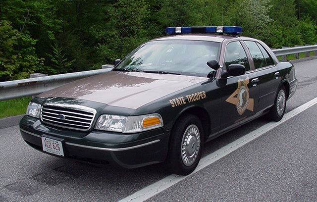Green and tan Crown Victoria State Trooper police car with blue light bar parked on highway shoulder