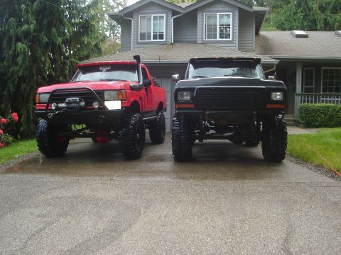 1989 Ford Bronco - Two heavily modified trucks, a red one with a snorkel and a black one, are parked side-by-side on a wet driveway.