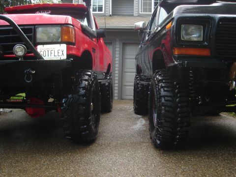 1989 Ford Bronco - Two heavily modified trucks, a red one and a black one, are parked in front of a garage.