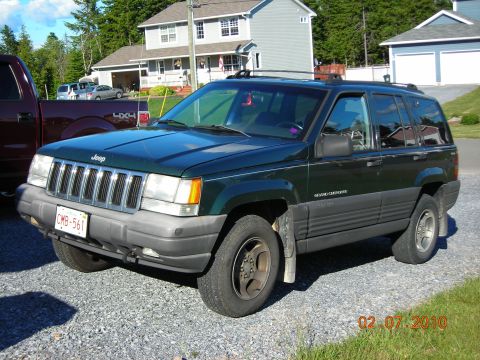 1984 Ford Bronco - A dark green Jeep Grand Cherokee is parked on a gravel driveway next to a maroon pickup truck.