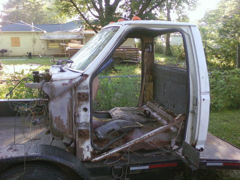 1989 Ford Bronco - A heavily weathered white truck cab sits on a flatbed trailer, stripped of its engine and doors.