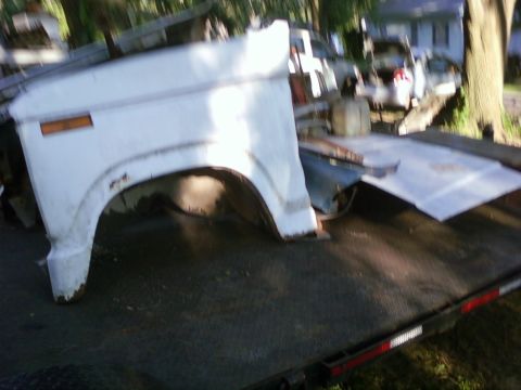 1989 Ford Bronco - A white truck fender and hood are loaded onto a flatbed trailer, with other vehicles and debris in the background.