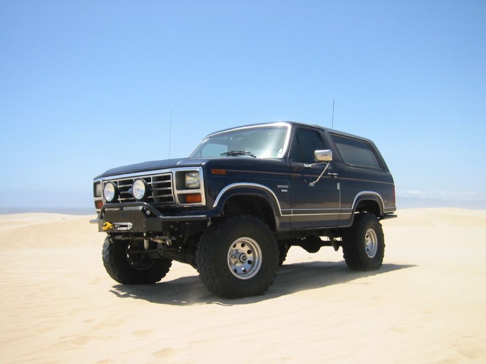 Lifted dark blue 1985 Ford Bronco with winch bumper and off-road tires on sand dunes