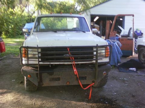 1989 Ford Bronco - Front view of a white pickup truck a bull bar and orange tie-down straps, with other vehicles and a garage in the background.