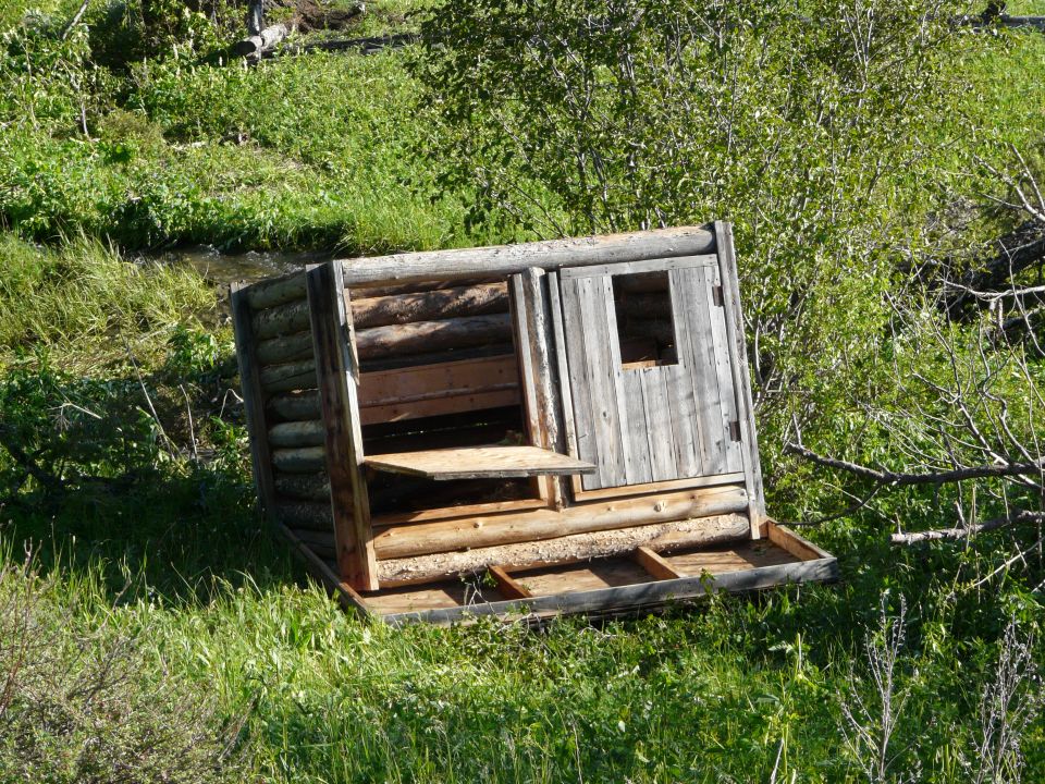 Wooden structure with a window and door, tilted on its side in grassy terrain