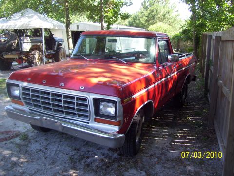1978 Ford Bronco - A red Ford pickup truck is parked next to a wooden fence, with another vehicle visible under a tent in the background.