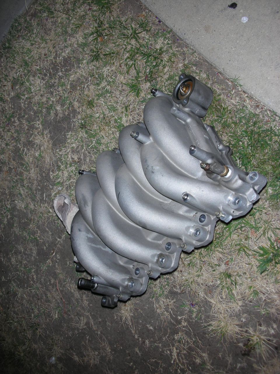 Aluminum intake manifold with multiple runners, resting on grass and concrete