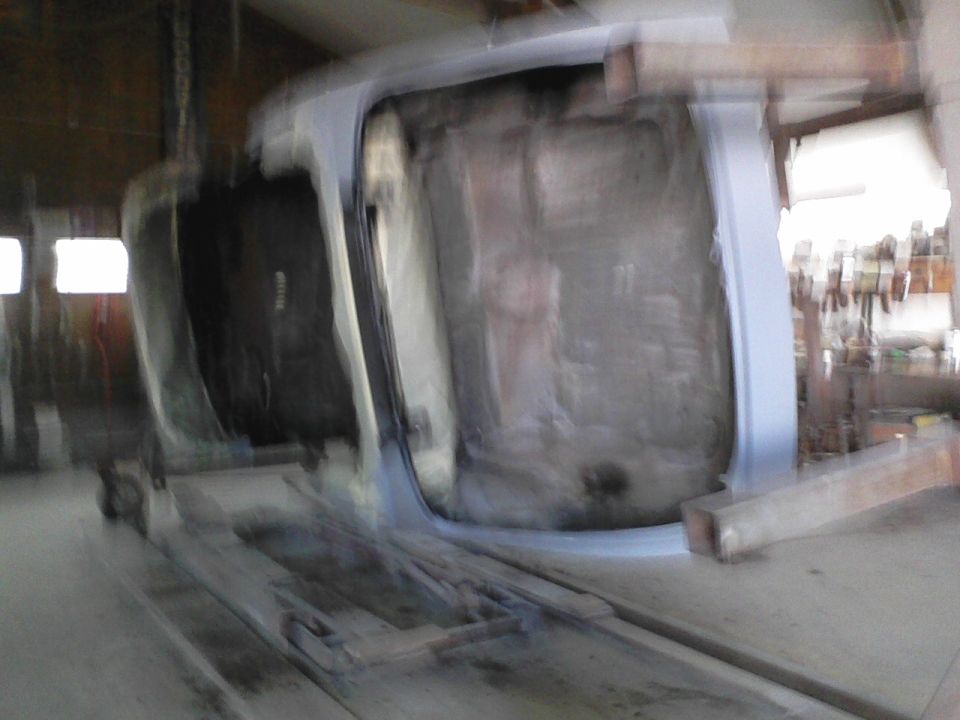 Car body upside down on a lift, showing the underside and interior. Some tools and shelves visible in background