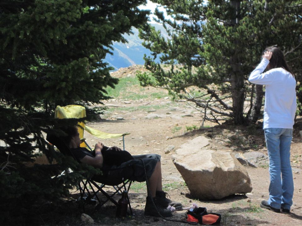 Two people in a wooded mountain area, one sitting in a chair, the other standing. A 'SUPER MOTORS' logo is visible at the bottom