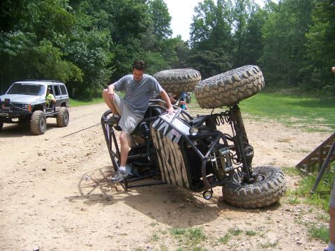 1992 Jeep YJ - An off-road buggy has tipped over on a dirt path, with a person perched on its frame while another vehicle stands by.