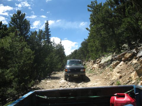 1978 Ford F-150 - A dark SUV navigates a rough, unpaved road surrounded by trees under a blue sky with clouds.