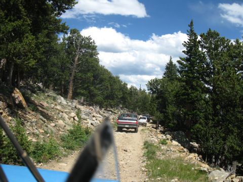 1978 Ford F-150 - A convoy of trucks, including a 1978 Ford F-150, navigates a rocky dirt path through a forested mountain landscape.