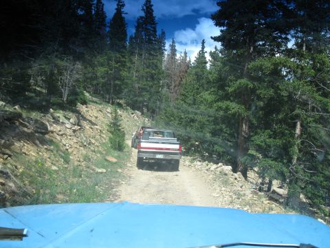 1978 Ford F-150 - Two pickup trucks navigate a dirt road through a dense forest with rocky terrain.
