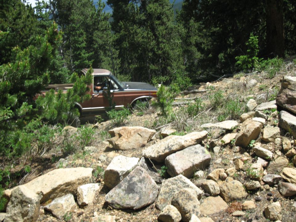 Brown pickup truck navigating a rough, rocky trail surrounded by trees. The truck appears to be from the late 1970s