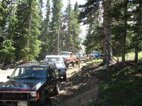 1978 Ford F-150 - A line of off-road vehicles navigates a wooded trail, with a Jeep Cherokee leading the pack.