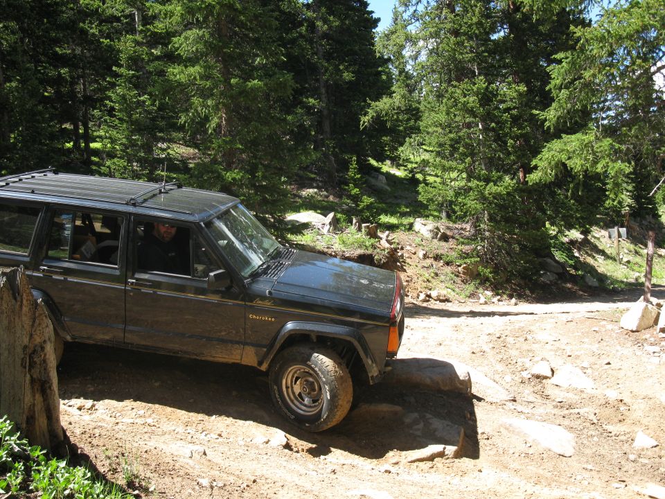 Dark-colored Jeep Cherokee on a dirt trail, driver visible, trees and rocks in background
