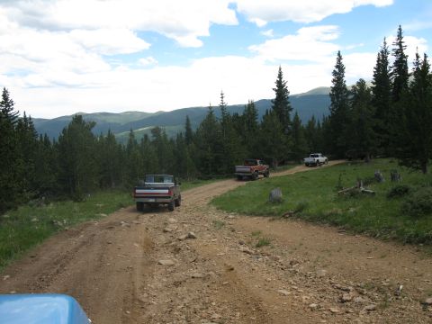 1978 Ford F-150 - Multiple pickup trucks navigate a winding dirt road in a scenic mountain landscape under a cloudy sky.