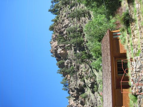 1978 Ford F-150 - A rustic wooden cabin is situated at the base of a steep, tree-covered cliff, with a bright blue sky overhead.