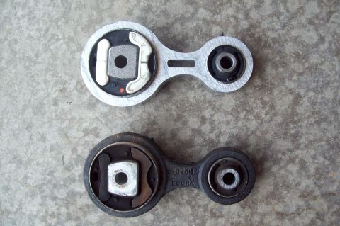 2006 Ford Fusion - Two engine/transmission torque strut mounts shown side by side on a concrete floor, one aluminum-bodied with visible rubber bushings and th…