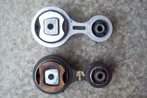 2006 Ford Fusion - Two lower engine/torque mounts (dogbone style) laid on a concrete floor, with the newer white-and-silver replacement on top showing part ma…