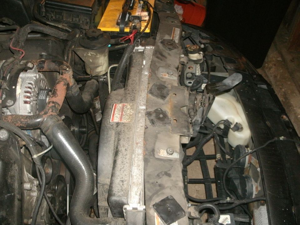 Car engine bay with radiator, alternator, hoses, and battery visible. Some labels are present