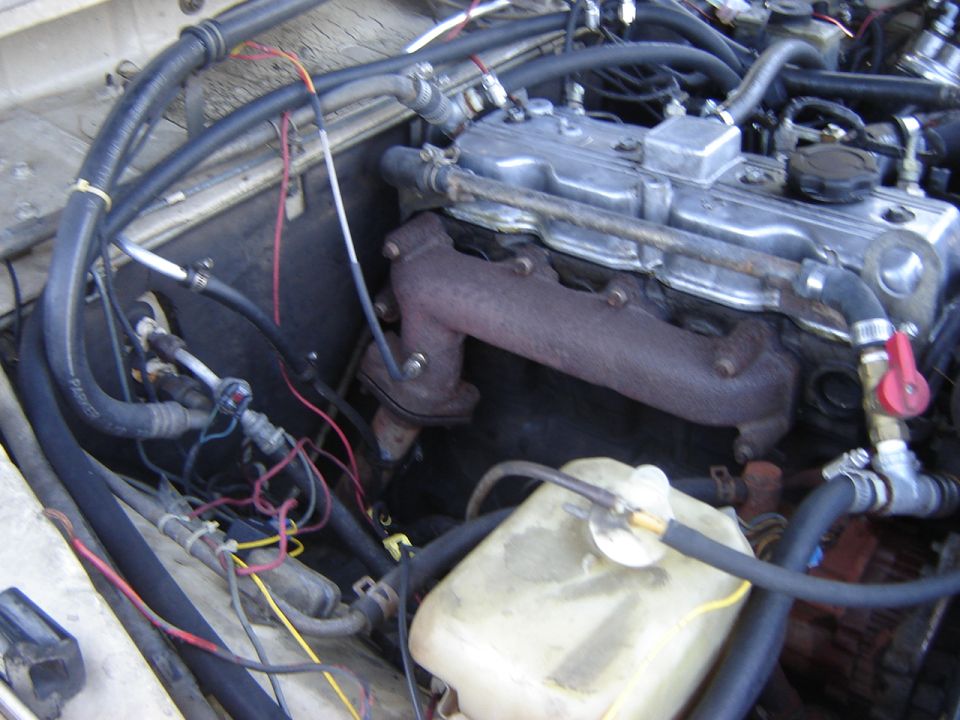 Engine bay with inline-four engine, rusty exhaust manifold, coolant overflow bottle, and various hoses and wiring