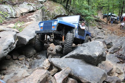1984 Ford Bronco II - A blue Jeep Wrangler tackles a challenging rocky trail, with other vehicles and spectators in the background. The Supermotors logo is visib…