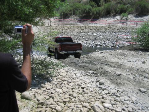 1986 Ford Bronco - A red pickup truck navigates a rocky stream bed, captured by a photographer in the foreground.