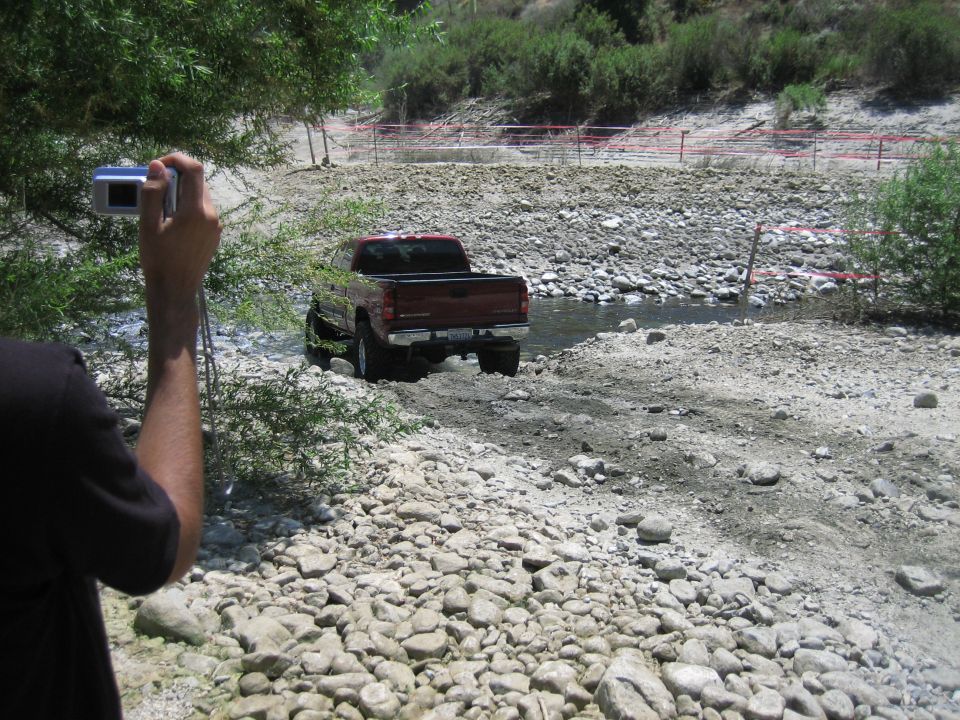 Red pickup truck driving through a rocky stream bed, with a person holding a camera in the foreground