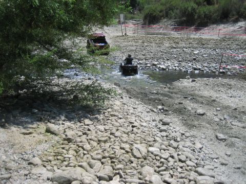 1986 Ford Bronco - Two off-road vehicles navigate a shallow, rocky stream under a tree's shade.