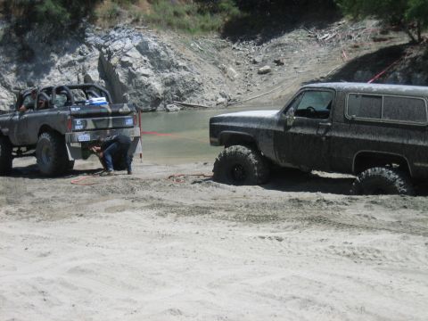 1986 Ford Bronco - A mud-splattered Chevrolet K5 Blazer is winched out of the sand by a modified pickup truck in a challenging off-road scenario.