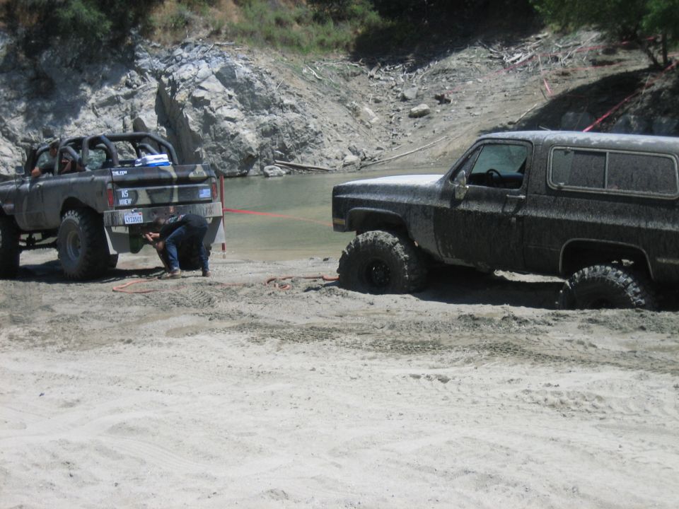 Mud-covered 1980s Chevrolet K5 Blazer stuck in sand, being winched by a modified pickup truck