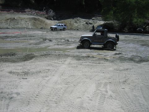 1986 Ford Bronco - Two off-road vehicles are seen in a muddy, sandy terrain with tire tracks visible.