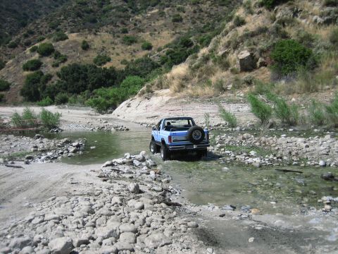 1986 Ford Bronco - A blue convertible SUV navigates a rocky stream in a dry, mountainous landscape.