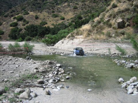 1986 Ford Bronco - A blue 1986 Ford Bronco navigates a rocky riverbed in a , natural landscape.