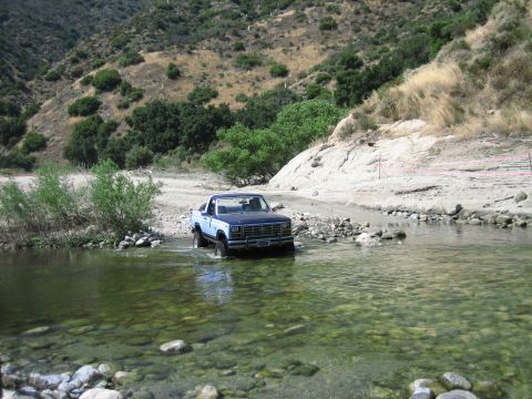 1986 Ford Bronco - A blue and white Ford pickup truck navigates a rocky stream bed in a dry, mountainous area.