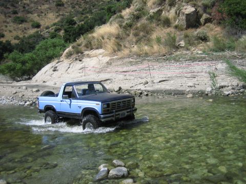 1986 Ford Bronco - A blue and black Ford Bronco navigates a clear, shallow river with rocky terrain.