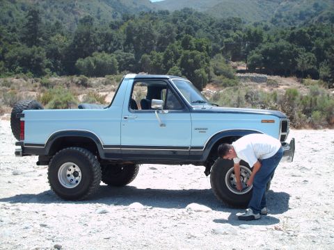 1986 Ford Bronco - A man inspects the front wheel of a light blue Ford Bronco parked on a dirt road with trees and hills in the background.