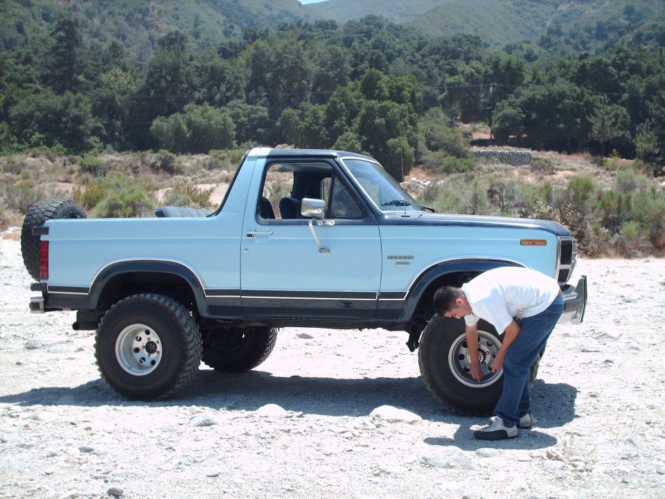 Man checking wheel on light blue 1986 Ford Bronco with spare tire on back