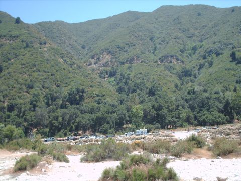 1986 Ford Bronco - A line of vehicles are parked in a dry, rocky area with a heavily wooded mountain range behind them.