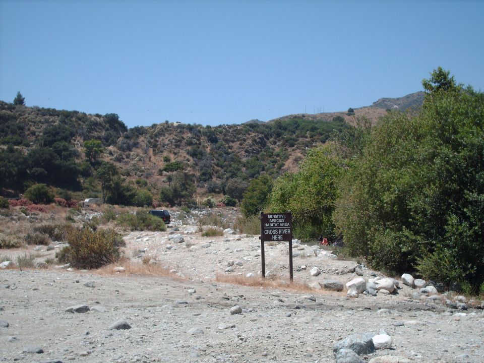 Dark SUV parked in dry riverbed with a "Cross River Here" sign