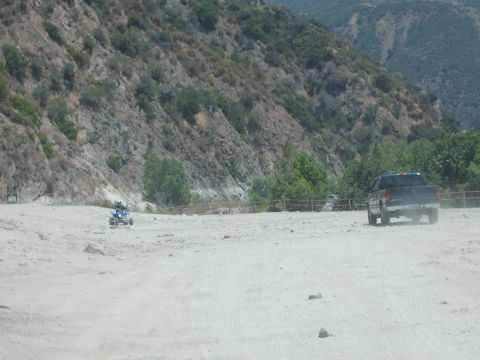 1986 Ford Bronco - A pickup truck kicks up dust on a dirt road, with an ATV visible in the background near a hillside.