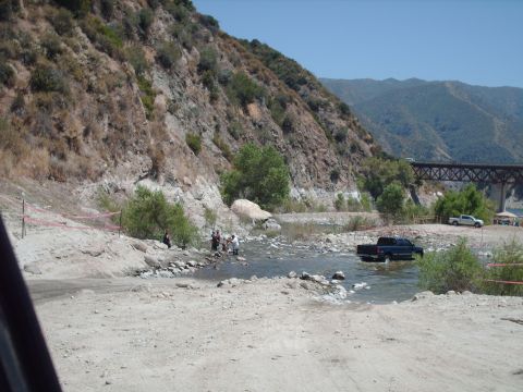1986 Ford Bronco - A dark pickup truck navigates a shallow river while a white pickup truck is parked on the shore, with a bridge visible in the distance.