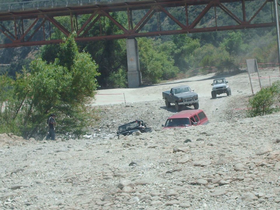 Several off-road vehicles, including a red SUV and a dark pickup truck, navigate a rocky, dry riverbed under a bridge