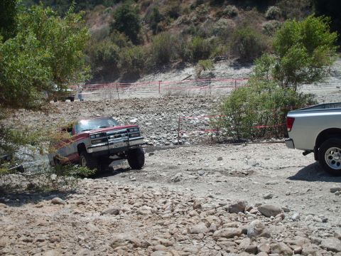 1986 Ford Bronco - A red and silver pickup truck navigates a rocky, off-road path, with another vehicle visible in the background.