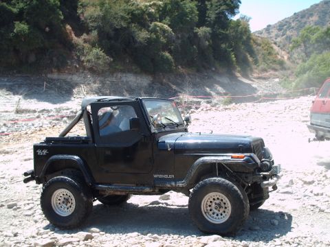 1986 Ford Bronco - A black Jeep Wrangler is shown off-road on a rocky terrain with brush and trees in the background.