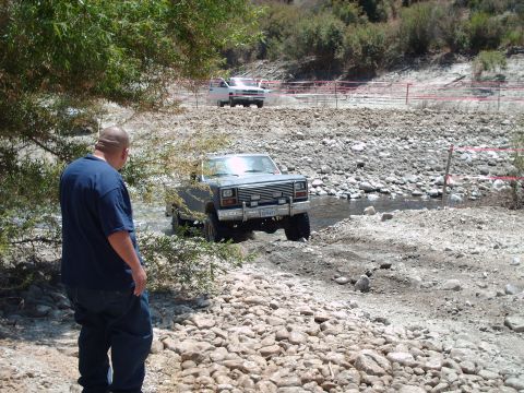 1986 Ford Bronco - A man watches a gray Ford Bronco navigate a rocky stream, with another white truck in the background.