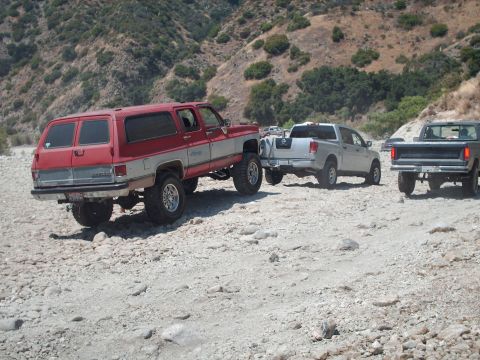 1986 Ford Bronco - Multiple trucks navigate a rocky terrain, with a lifted red and silver SUV being towed by a silver pickup.
