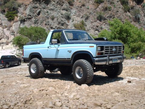 1986 Ford Bronco - This lifted 1980 Ford Bronco features a two-tone blue paint scheme and off-road tires, parked in a rocky, outdoor setting.