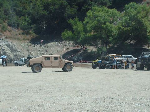 1986 Ford Bronco - Several off-road vehicles are gathered in a dusty lot with a wooded hillside in the background.